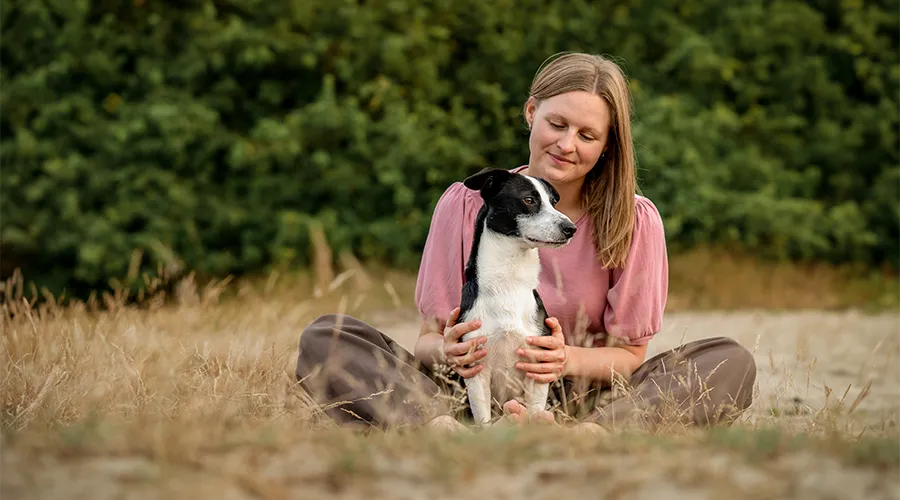 Frau sitzt im Schneidersitz im Dünengras, ihr kleiner schwarz-weißer Hund lehnt an ihrer Schulter, Hundefotografie in Bremen und Niedersachsen von Ines Arnshoff.