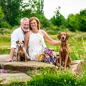 Glückliches Paar sitzt nach seinem Fotoshooting mit Hund in einer blühenden Wiese in Weyhe Niedersachsen, daneben seine zwei Hunde — ein Magyar Vizsla und ein kleiner Terrier, Hundefotografie in Niedersachsen von Ines Arnshoff