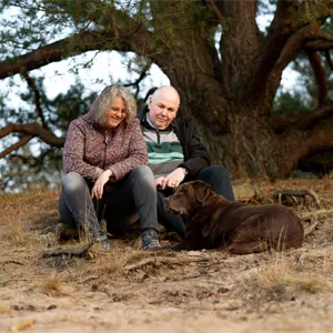 Reifes Paar sitzt unter einer Kiefer in der Heide, davor liegt entspannt sein einäugiger schokoladenbrauner Labrador, Hundefotografie in Bremen von Ines Arnshoff