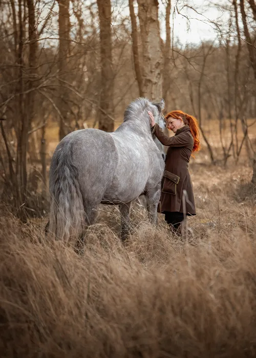 Rothaarige Frau lehnt ihre Stirn zärtlich an den Kopf ihres Apfelschimmels im winterlichen Birkenwald, Tierfotografie in Bremen und Niedersachsen von Ines Arnshoff.