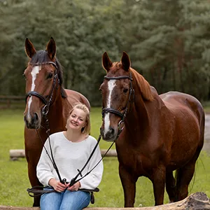 Blonde Kundin sitzt bei ihrem Fotoshooting mit zwei Pferden auf einem Baumstamm und blickt lächelnd zu ihren beiden Warmblütern auf, Pferdefotografie in Bremen und Niedersachsen von Ines Arnshoff