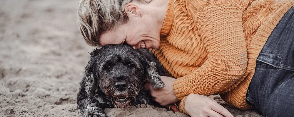 Tierfotografin Ines Arnshoff aus Bremen liegt lachend im Sand, die Wange an den Kopf ihres Seelenhunds Winnie gelehnt, einer schwarzen Portugiesischen Wasserhündin.