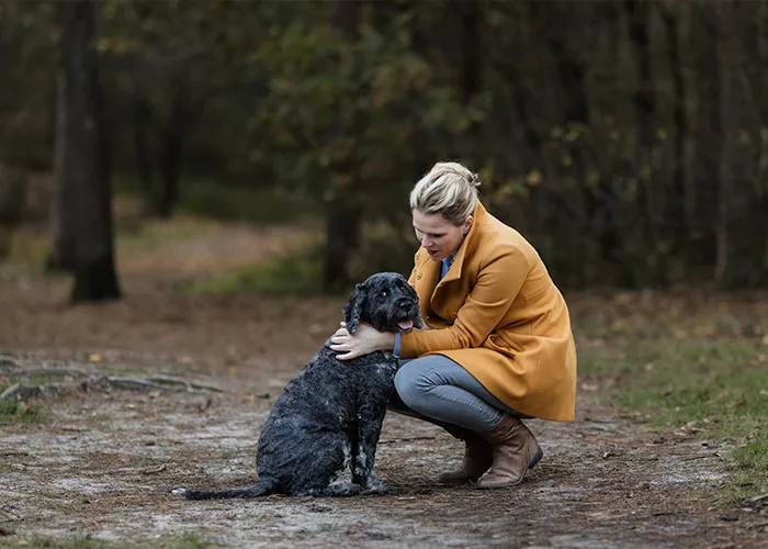 Ines Arnshoff geht im Herbstwald in die Hocke und legt die Hand auf den Rücken ihrer schwarzen Hündin Winnie.