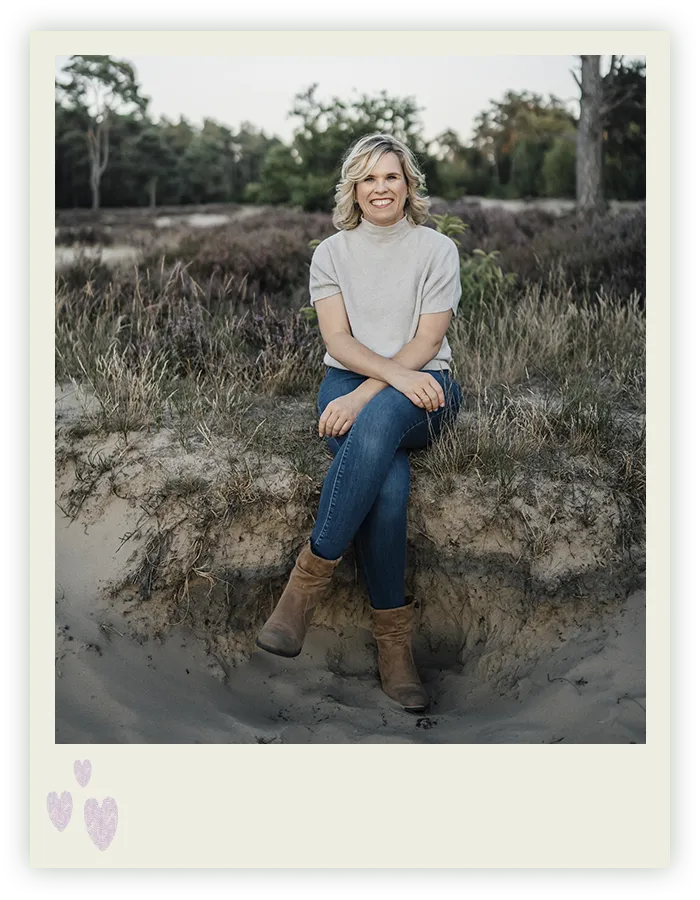 Ines Arnshoff sitzt lächelnd mit übergeschlagenen Beinen auf einer Sandkante in der Heidelandschaft